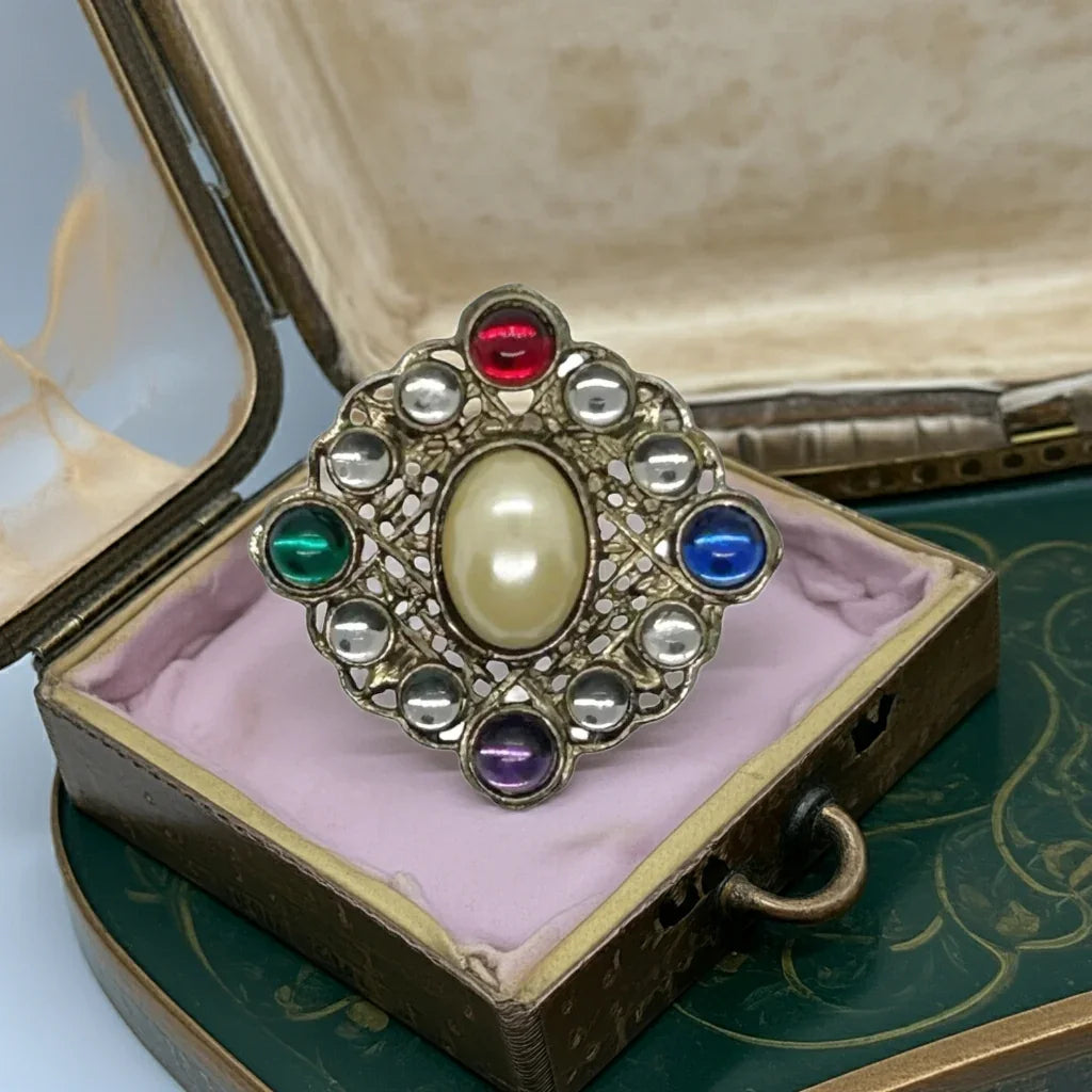 This striking brooch boasts a majestic diamond design, crafted from intricately detailed filigree metal and adorned with a sizable oval faux pearl at its heart. Surrounding the pin are lustrous round cabochons in a rich palette of red, green, purple, and blue, elegantly interspersed with two clear gems between each color