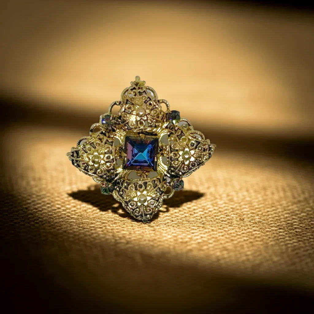 This exquisite brooch with an Old World aesthetic showcases finely crafted gold filigree petals encircling a square stone in elegant blue-purple tones. Between each delicate petal rests a round, iridescent stone that complements the central hues, adding subtle depth and shimmer. 