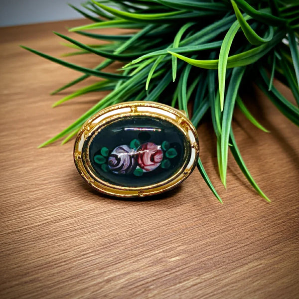 Picture a charming piece of wearable nostalgia: this oval brooch pairs a moody black canvas with cheerful painted florals, all framed in warm gold-tone metal. It's a mid-20th century take on Victorian mourning jewelry aesthetics