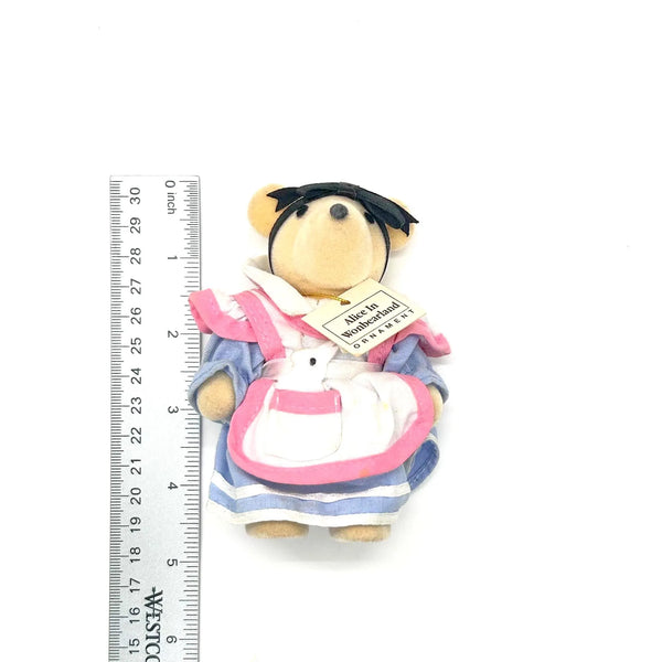 A small plush teddy bear dressed in a pink and blue apron with a white pocket, standing next to a metal ruler on a white background. The tag reads 'All in All out - Toy Store'.