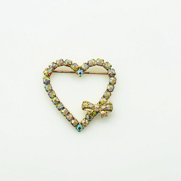 This charming mid-century brooch captures all the sparkle and romance of vintage glamour. The delicate gold-tone heart showcases shimmering aurora borealis rhinestones that dance with rainbow fire, while a sweet bow accent adds an extra touch of whimsy.