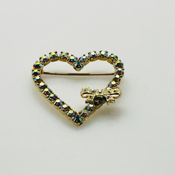 This charming mid-century brooch captures all the sparkle and romance of vintage glamour. The delicate gold-tone heart showcases shimmering aurora borealis rhinestones that dance with rainbow fire, while a sweet bow accent adds an extra touch of whimsy.