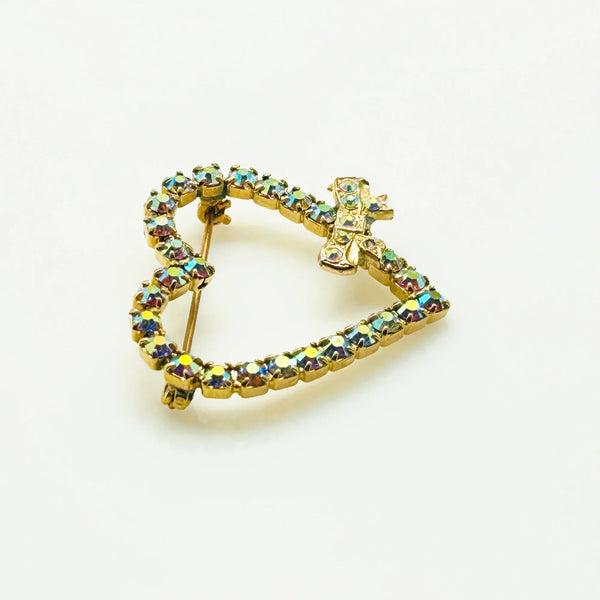 This charming mid-century brooch captures all the sparkle and romance of vintage glamour. The delicate gold-tone heart showcases shimmering aurora borealis rhinestones that dance with rainbow fire, while a sweet bow accent adds an extra touch of whimsy.