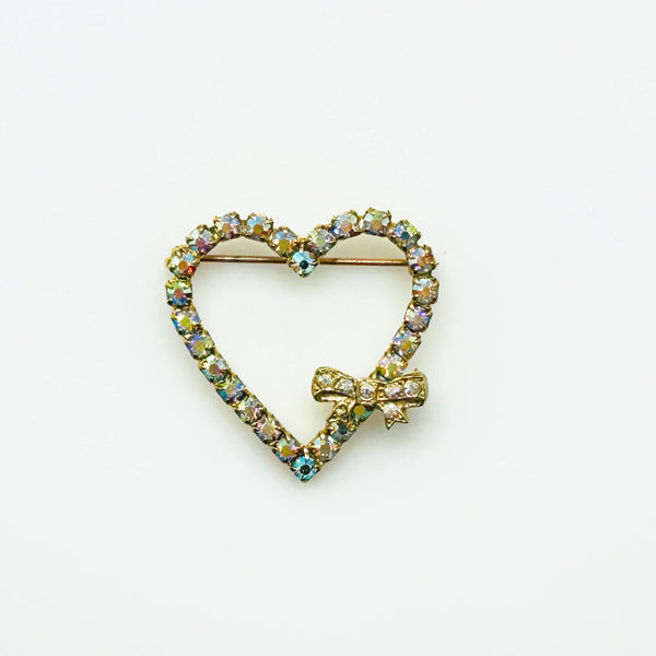 This charming mid-century brooch captures all the sparkle and romance of vintage glamour. The delicate gold-tone heart showcases shimmering aurora borealis rhinestones that dance with rainbow fire, while a sweet bow accent adds an extra touch of whimsy.
