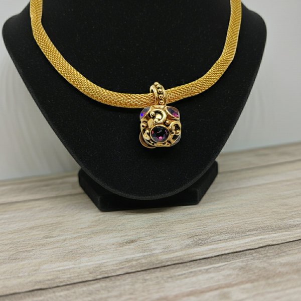 This 1990s pendant necklace features a meticulously crafted design with round purple faceted gemstones set in bezel-mounted antiqued gold metalwork. The ornamental pendant is suspended from a rounded mesh chain, measuring 16" secured with a toggle clasp.