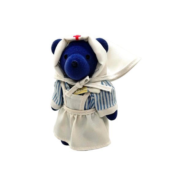 A small plush teddy bear dressed as a nurse, with a blue body, white apron and head covering, and a small red cross on the hat, photographed on a clean white background.