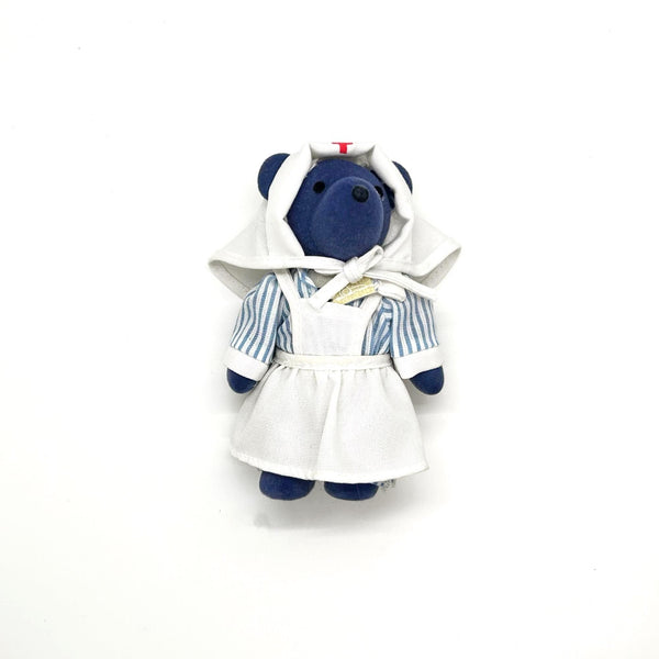 A small plush teddy bear dressed as a nurse, featuring a navy blue body, a white nurse hat with a tiny red cross, and a blue-and-white striped outfit, set against a plain white background.