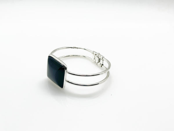 This striking vintage bracelet showcases a bold black square onyx stone set in polished silver. The architectural design features dual silver bands converging at the center stone with a hinged hook-bangle closure for easy wear. 