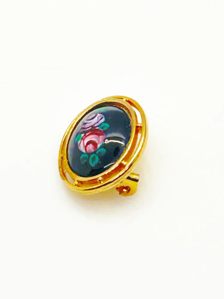 Picture a charming piece of wearable nostalgia: this oval brooch pairs a moody black canvas with cheerful painted florals, all framed in warm gold-tone metal. It's a mid-20th century take on Victorian mourning jewelry aesthetics