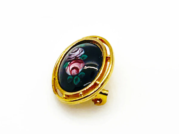 Picture a charming piece of wearable nostalgia: this oval brooch pairs a moody black canvas with cheerful painted florals, all framed in warm gold-tone metal. It's a mid-20th century take on Victorian mourning jewelry aesthetics