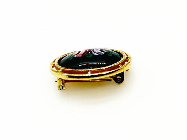 Picture a charming piece of wearable nostalgia: this oval brooch pairs a moody black canvas with cheerful painted florals, all framed in warm gold-tone metal. It's a mid-20th century take on Victorian mourning jewelry aesthetics