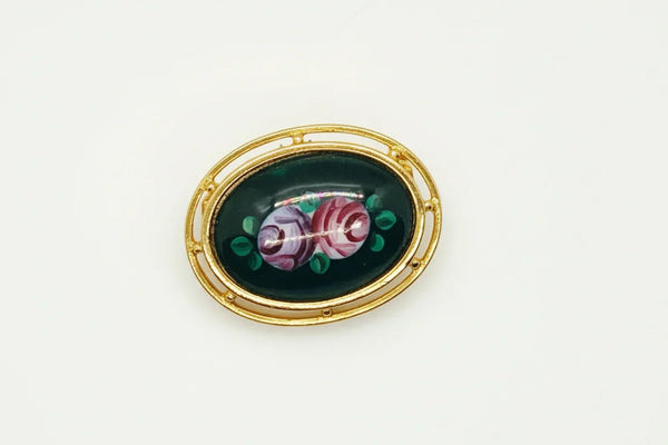 Picture a charming piece of wearable nostalgia: this oval brooch pairs a moody black canvas with cheerful painted florals, all framed in warm gold-tone metal. It's a mid-20th century take on Victorian mourning jewelry aesthetics