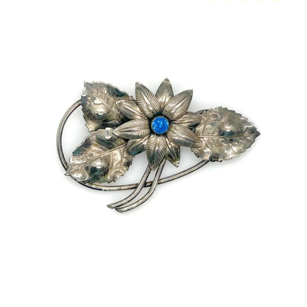 A vintage silver-tone floral brooch featuring a central flower with a blue cabochon stone, surrounded by textured leaves and stems, set against a plain white background.