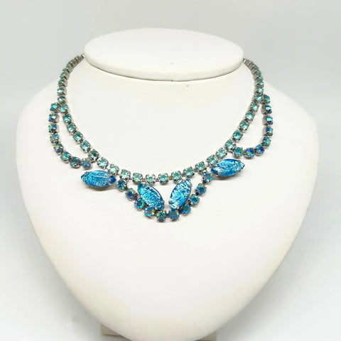This stunning vintage necklace features a simple chain of medium blue rhinestones. The centerpiece is truly captivating, with three elements—each made of a strand of blue rhinestones—draping over marquise-shaped, iridescent blue confetti lucite. 