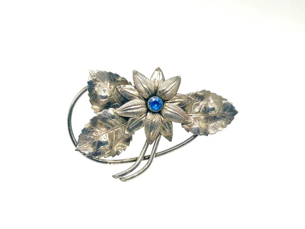 A vintage-style silver brooch featuring a flower motif with textured leaves and a central round blue gemstone, set against a plain white background.