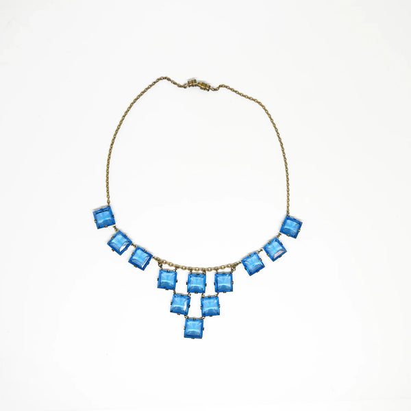 Experience the height of sophistication with this exquisite vintage Art Deco necklace. Featuring a flawless line of rich azure square stones, delicately connected by two additional rows, all set on a luxurious gold tone link chain. The stones are meticulously held in place with thin claw settings, revealing their exposed backs for an added touch of elegance.