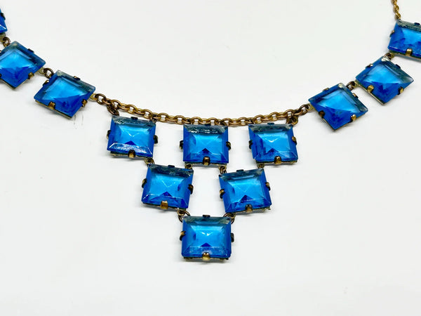 Experience the height of sophistication with this exquisite vintage Art Deco necklace. Featuring a flawless line of rich azure square stones, delicately connected by two additional rows, all set on a luxurious gold tone link chain. The stones are meticulously held in place with thin claw settings, revealing their exposed backs for an added touch of elegance.