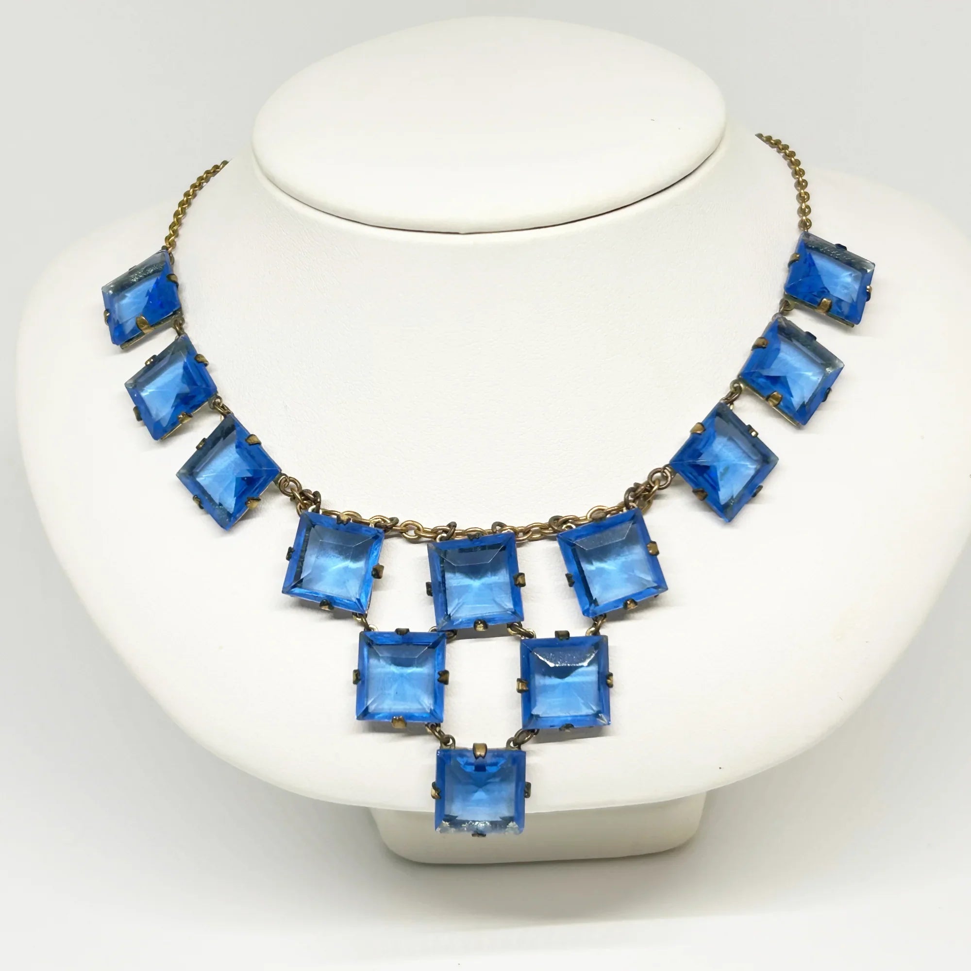 Experience the height of sophistication with this exquisite vintage Art Deco necklace. Featuring a flawless line of rich azure square stones, delicately connected by two additional rows, all set on a luxurious gold tone link chain. The stones are meticulously held in place with thin claw settings, revealing their exposed backs for an added touch of elegance.
