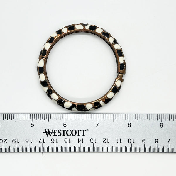 This striking hinge bangle bracelet showcases bold geometric patterns rendered in contrasting black and white enamel, accented with warm copper metalwork. The repeating abstract design creates visual interest from every angle, making it a versatile piece that complements both casual and dressed-up looks