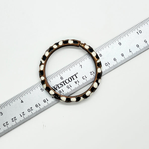 This striking hinge bangle bracelet showcases bold geometric patterns rendered in contrasting black and white enamel, accented with warm copper metalwork. The repeating abstract design creates visual interest from every angle, making it a versatile piece that complements both casual and dressed-up looks