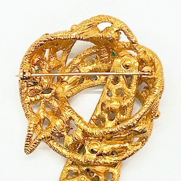 An image showing the reverse side of a textured gold-tone vintage costume jewelry brooch. The brooch is shaped like a textured, openwork abstract ribbon loop or bow with a dangling end pointing downward. A long, horizontal metal pin stem and safety clasp run across the upper back portion. On the lower section of the brooch, an oval cartouche containing the Capri maker's mark is stamped directly onto the textured filigree-style metal framework
