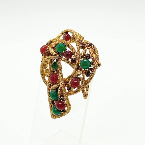 An image showing the reverse side of a textured gold-tone vintage costume jewelry brooch. The brooch is shaped like a textured, openwork abstract ribbon loop or bow with a dangling end pointing downward. A long, horizontal metal pin stem and safety clasp run across the upper back portion. On the lower section of the brooch, an oval cartouche containing the Capri maker's mark is stamped directly onto the textured filigree-style metal framework