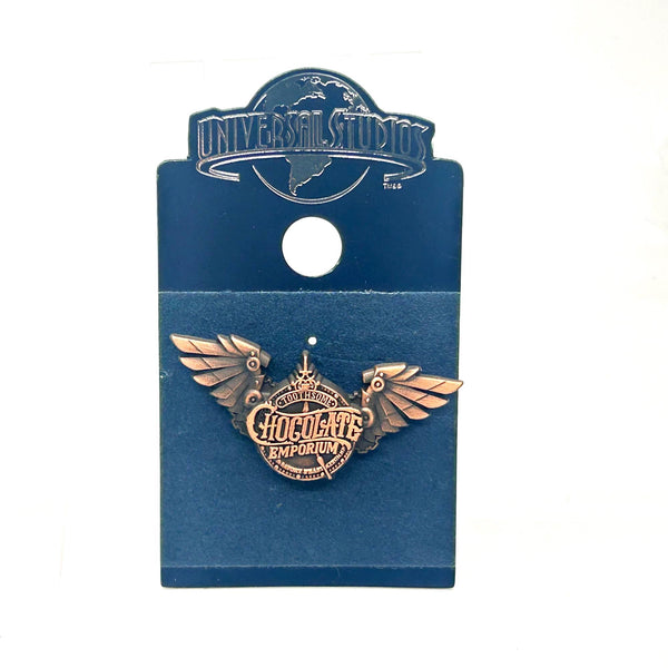 Official Universal Studios Parks Toothsome Chocolate Emporium steampunk logo lapel pin with mechanical wings, mounted on its original dark blue backing card.