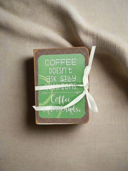 Handcrafted miniature book with a brown rustic border, a sage green front cover, and a white ribbon tied around the center. The cover features a white hand-lettered quote that reads, "Coffee doesn't ask silly questions. Coffee understands." 