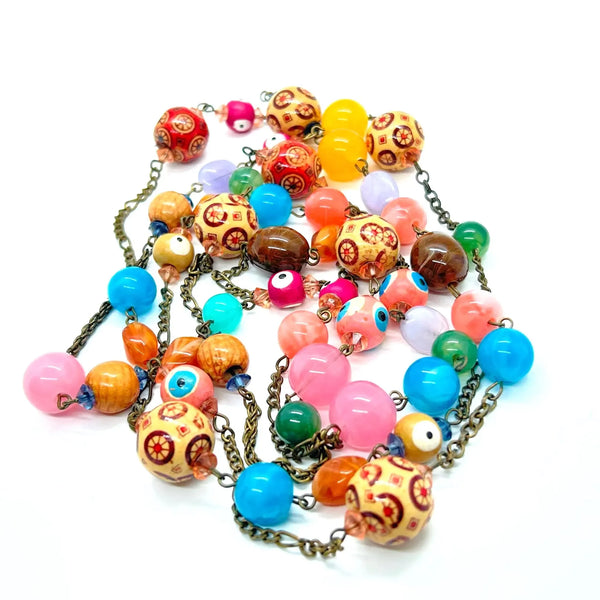 A colorful beaded necklace with a mix of pink, blue, orange, yellow, green, and brown beads, including evil eye and patterned round beads, attached to an antique bronze chain on a white background.