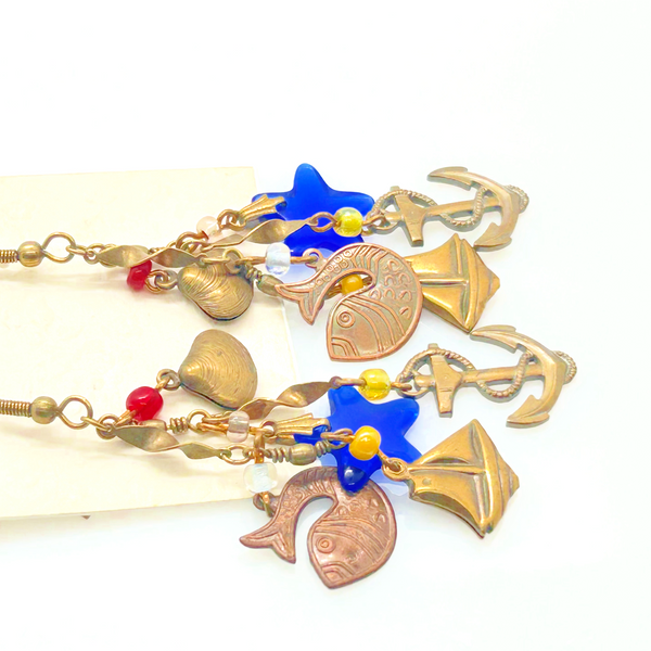 Vintage Cookie Lee nautical charm dangle earrings in antiqued brass with anchor, sailboat, fish, and blue star charms on a cream card.