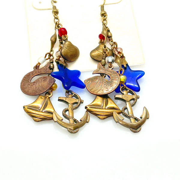 Vintage Cookie Lee nautical charm dangle earrings in antiqued brass with anchor, sailboat, fish, and blue star charms on a cream card.