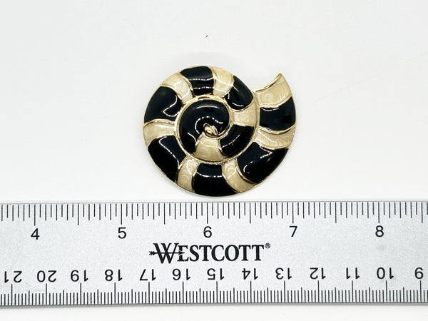 This stunning Trifari brooch captures the elegance of Art Deco design with its spiral seashell silhouette. Crafted in gold-tone metal, it features striking black enamel paired with creamy golden-sand enamel in a sophisticated moire finish.