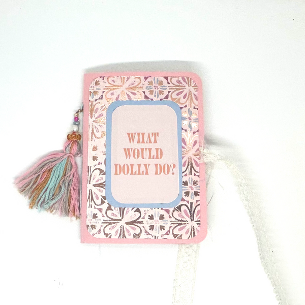 A small handmade journal or notebook with a pink border, floral patterned cover, and a tassel on the left side. The center features a light blue rounded rectangle with the text 'WHAT WOULD DOLLY DO?' in pink.