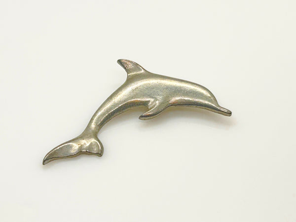A vintage pewter dolphin brooch shown flat against a plain, off-white background. The brooch features a minimalist, sleek silver-tone design capturing a dolphin in a leaping arc, with a smooth matte finish, structured dorsal fin, flippers, and a detailed tail flipper.