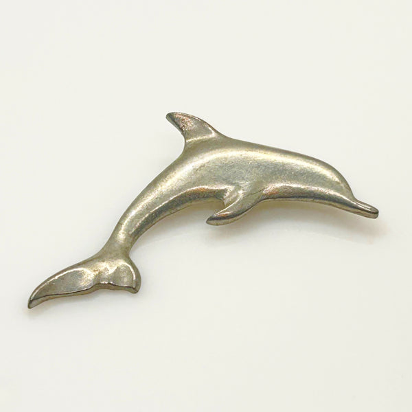 A vintage pewter dolphin brooch shown flat against a plain, off-white background. The brooch features a minimalist, sleek silver-tone design capturing a dolphin in a leaping arc, with a smooth matte finish, structured dorsal fin, flippers, and a detailed tail flipper.