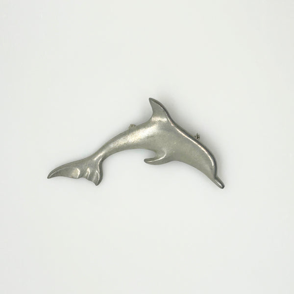 A vintage pewter dolphin brooch shown flat against a plain, off-white background. The brooch features a minimalist, sleek silver-tone design capturing a dolphin in a leaping arc, with a smooth matte finish, structured dorsal fin, flippers, and a detailed tail flipper.