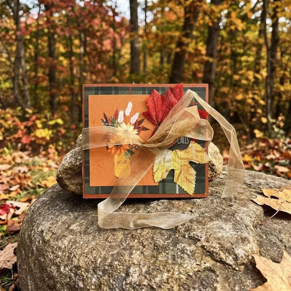 Handcrafted fall mini scrapbook cover featuring a green and black plaid background with an orange center panel. The design is decorated with a paper leaf printed with text, a dark red faux maple leaf, and a small autumn floral arrangement. A wide, sheer gold organza ribbon is tied into a large bow across the center
