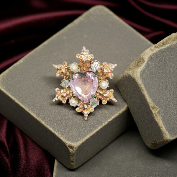 A gold-toned brooch with a central heart-shaped, light purple gemstone. The brooch is surrounded by ornate gold filigree, small pearls, and clear crystals, creating a vintage, elegant design.
