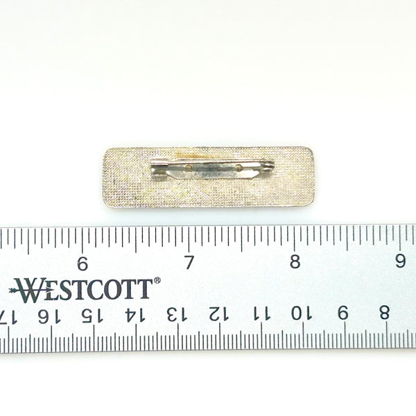 A vintage rectangular Art Deco revival bar brooch in the Pierre Bex style, featuring textured gold-tone metal sunburst rays, dark enamel details, and a central floral cloud medallion against a solid white background