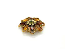 A 1950s Austrian costume jewelry flower pin featuring tiered, multi-colored faceted crystals in warm amber, yellow, and brown tones. The center cluster is arranged with small, round olivine green rhinestones, set on a gold-tone filigree metal backing against a plain white background.