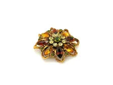 A 1950s Austrian costume jewelry flower pin featuring tiered, multi-colored faceted crystals in warm amber, yellow, and brown tones. The center cluster is arranged with small, round olivine green rhinestones, set on a gold-tone filigree metal backing against a plain white background.