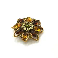 A 1950s Austrian costume jewelry flower pin featuring tiered, multi-colored faceted crystals in warm amber, yellow, and brown tones. The center cluster is arranged with small, round olivine green rhinestones, set on a gold-tone filigree metal backing against a plain white background.