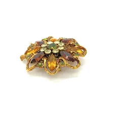 A 1950s Austrian costume jewelry flower pin featuring tiered, multi-colored faceted crystals in warm amber, yellow, and brown tones. The center cluster is arranged with small, round olivine green rhinestones, set on a gold-tone filigree metal backing against a plain white background.