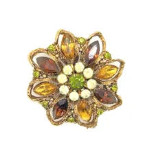 A 1950s Austrian costume jewelry flower pin featuring tiered, multi-colored faceted crystals in warm amber, yellow, and brown tones. The center cluster is arranged with small, round olivine green rhinestones, set on a gold-tone filigree metal backing against a plain white background.