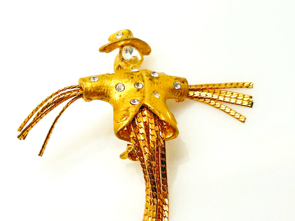 Detailed close-up of a vintage figural scarecrow brooch showing the textured metal patterns, embedded clear chaton rhinestones, and individual strands of fluid herringbone chain hanging from the tors