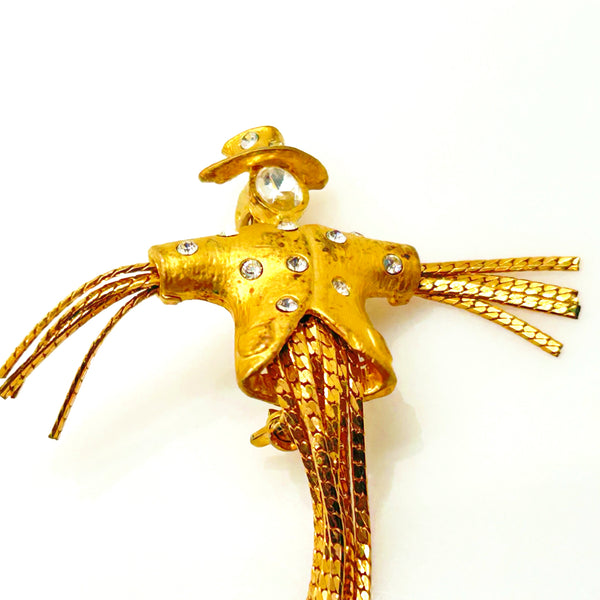 Detailed close-up of a vintage figural scarecrow brooch showing the textured metal patterns, embedded clear chaton rhinestones, and individual strands of fluid herringbone chain hanging from the tors