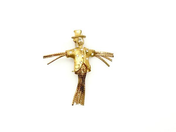 Vintage gold-tone scarecrow brooch pin with a textured finish, clear rhinestone accents on the coat and hat, and a lower body made of swinging gold chain fringe to look like straw legs, set against a plain white background