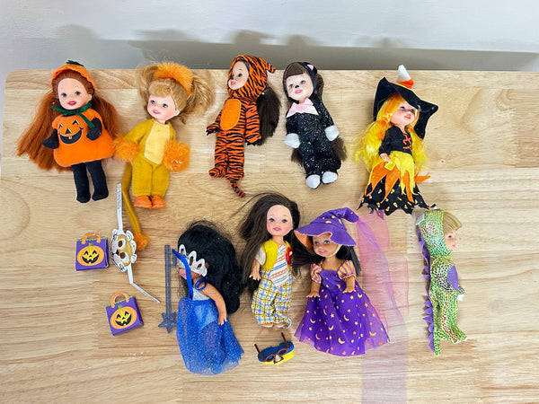 A collection of eight vintage Mattel Barbie Kelly Club mini dolls in Halloween Party costumes arranged on a light wood surface. The assortment features dolls dressed as a pumpkin with a Jack-o'-lantern trick-or-treat bag, a tiger, a black cat, witches with pointed hats, a mermaid in a blue glitter dress, and a green creature, alongside small plastic Halloween accessories.
