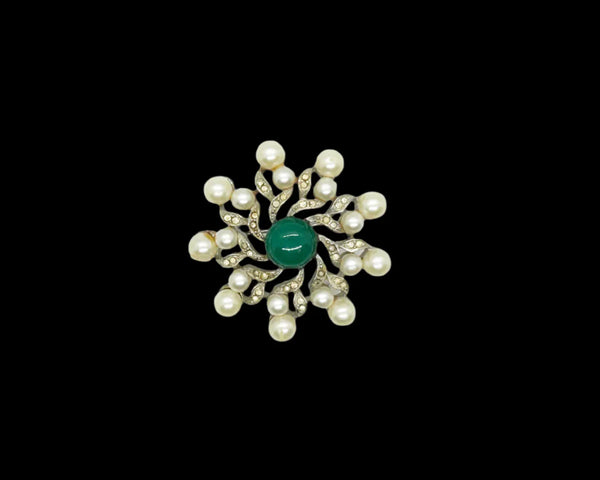  Radiating a regal charm reminiscent of a treasured royal heirloom, it captivates with its intricate design and flawless condition. Measuring 1 3/4" wide, the brooch features a striking green round cabochon centerpiece surrounded by elegant swirling patterns. Cream faux pearls add a soft, classic touch, all set in a lustrous silver-tone metal that enhances its vintage appeal.