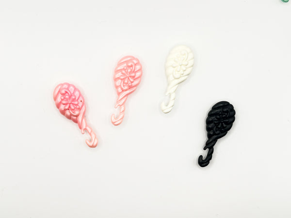 A collection of three miniature plastic doll styling accessories from the 1980 Mattel Western Barbie line. The lot includes a metallic teal green brush, a pale pastel pink brush, and a vibrant hot pink brush. Each item is molded with an intricate, traditional floral pattern on the back and ends in a curved hook-style clip handle meant to clip onto a doll's hand
